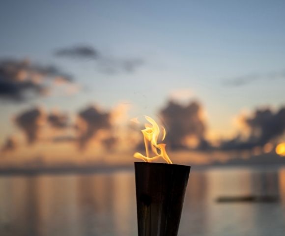 A lit torch against a calm sea at sunset, reflecting vibrant orange and blue hues. The scene exudes serenity and warmth, with soft clouds in the sky.
