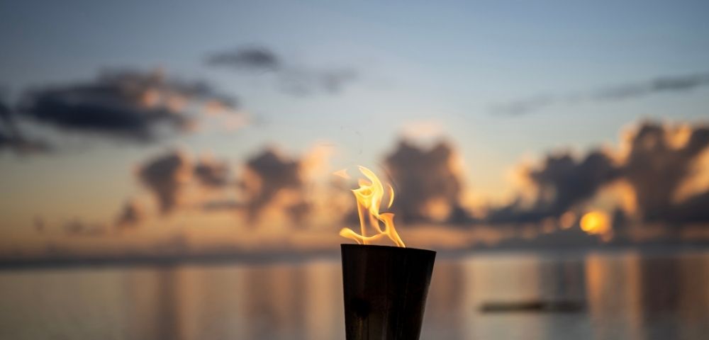 A lit torch against a calm sea at sunset, reflecting vibrant orange and blue hues. The scene exudes serenity and warmth, with soft clouds in the sky.
