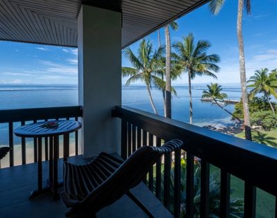 Balcony overlooking a serene beach with palm trees under a clear blue sky. Two wooden chairs and a small table create a peaceful, tropical vibe.