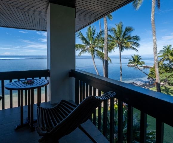 Balcony overlooking a serene beach with palm trees under a clear blue sky. Two wooden chairs and a small table create a peaceful, tropical vibe.
