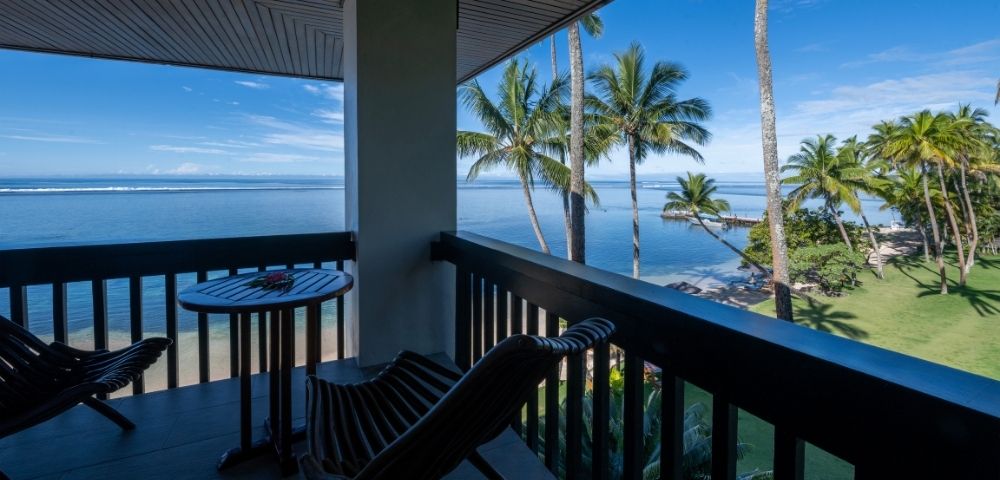 Balcony overlooking a serene beach with palm trees under a clear blue sky. Two wooden chairs and a small table create a peaceful, tropical vibe.