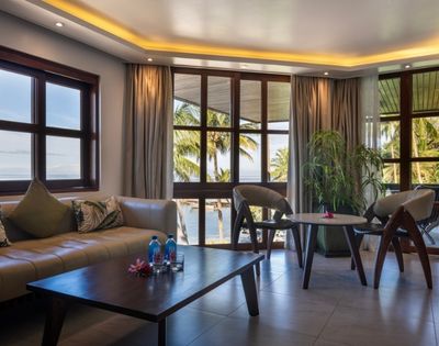 A serene living room with large windows overlooking palm trees and water. It has a beige sofa, two chairs, a wooden table, and soft lighting.