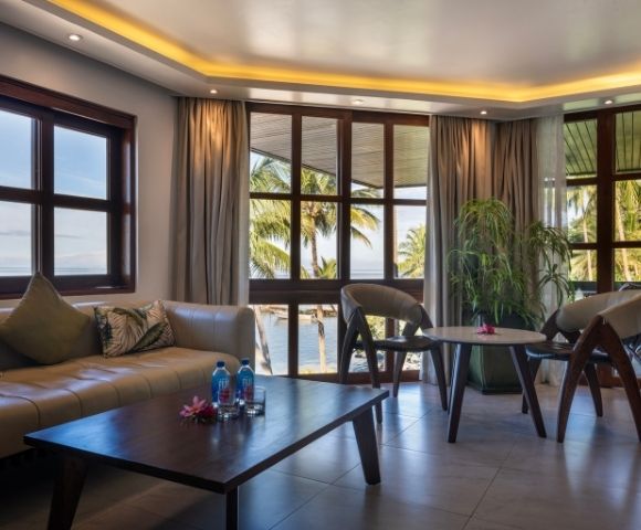 A serene living room with large windows overlooking palm trees and water. It has a beige sofa, two chairs, a wooden table, and soft lighting.