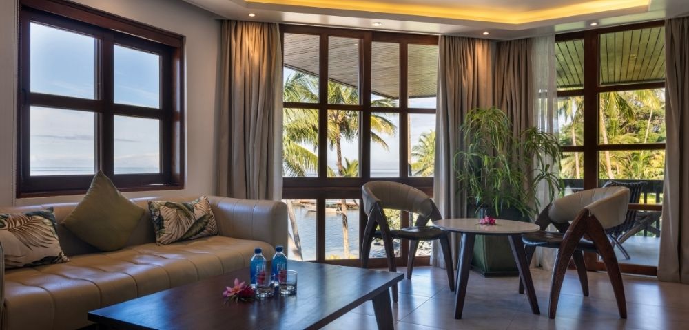 A serene living room with large windows overlooking palm trees and water. It has a beige sofa, two chairs, a wooden table, and soft lighting.