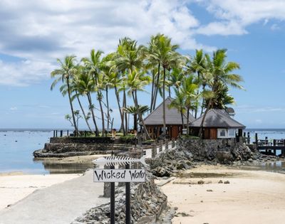 Seaside tropical scene with palm trees, a thatched-roof hut, and a sandy beach. A sign reads 