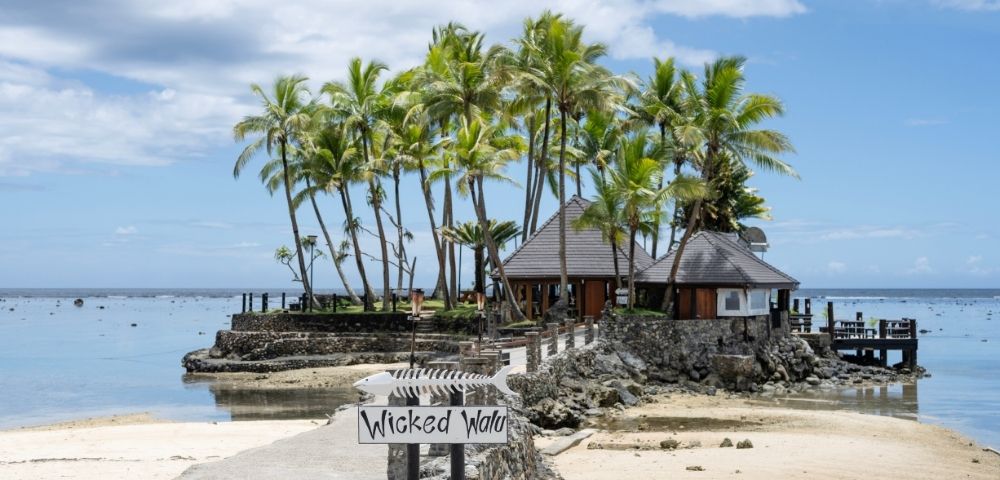 Seaside tropical scene with palm trees, a thatched-roof hut, and a sandy beach. A sign reads 