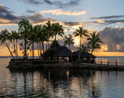 Tropical island with palm trees at sunset, silhouetted against an orange sky with scattered clouds. Calm water reflects the serene scene. Peaceful ambiance.