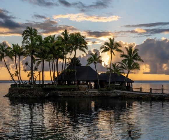 Tropical island with palm trees at sunset, silhouetted against an orange sky with scattered clouds. Calm water reflects the serene scene. Peaceful ambiance.