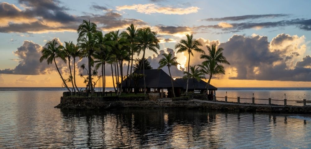 Tropical island with palm trees at sunset, silhouetted against an orange sky with scattered clouds. Calm water reflects the serene scene. Peaceful ambiance.
