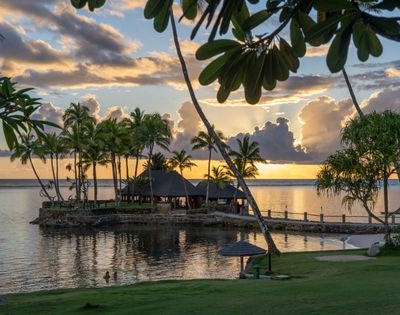 Tropical island scene at sunset with silhouetted palm trees by calm water, vibrant orange and yellow sky, and a peaceful, relaxed atmosphere.