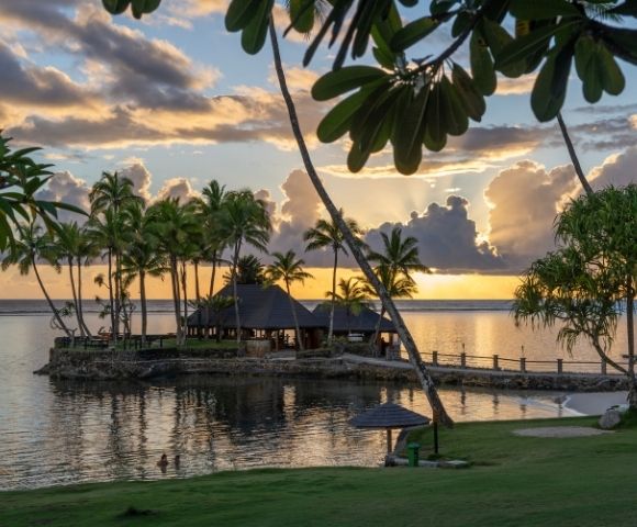 Tropical island scene at sunset with silhouetted palm trees by calm water, vibrant orange and yellow sky, and a peaceful, relaxed atmosphere.