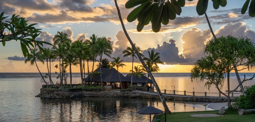 Tropical island scene at sunset with silhouetted palm trees by calm water, vibrant orange and yellow sky, and a peaceful, relaxed atmosphere.