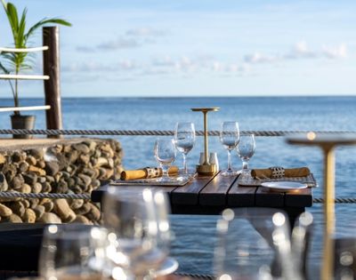 Elegant seaside dining scene with a wooden table set for four, featuring wine glasses, napkins, and a candleholder. The ocean view is serene and inviting.
