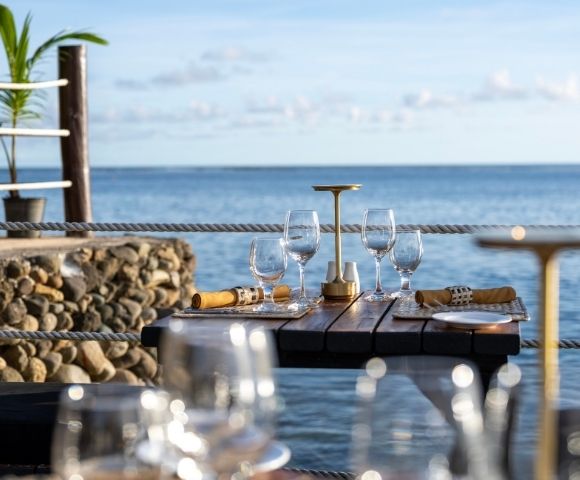Elegant seaside dining scene with a wooden table set for four, featuring wine glasses, napkins, and a candleholder. The ocean view is serene and inviting.