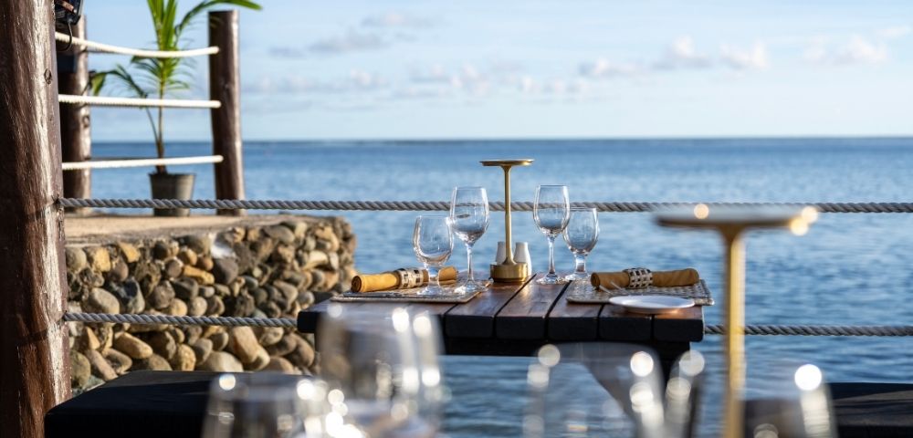 Elegant seaside dining scene with a wooden table set for four, featuring wine glasses, napkins, and a candleholder. The ocean view is serene and inviting.