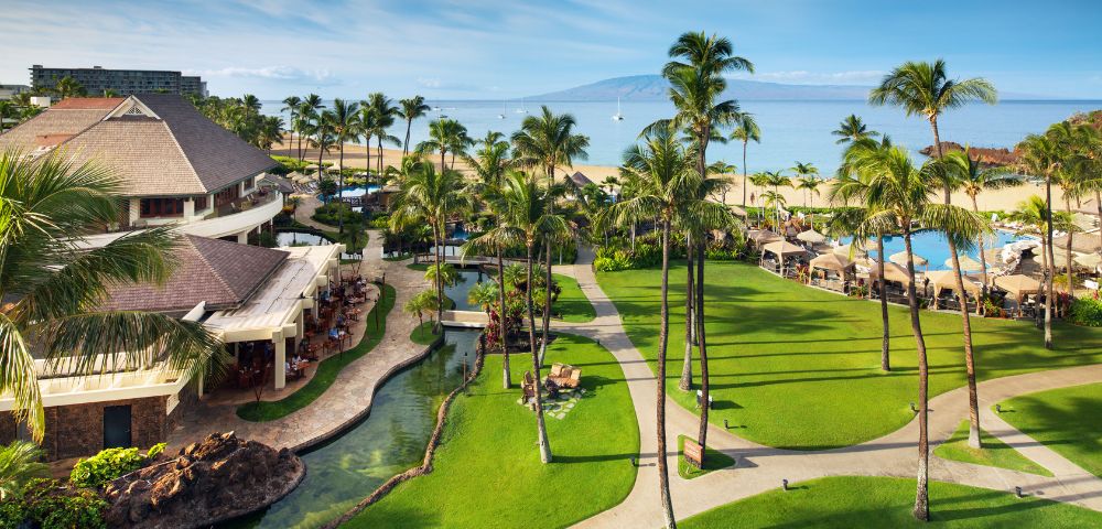Tropical resort view with lush green lawns, palm trees, and a curved pathway. A pool and cabanas are near a sandy beach with calm ocean waves.