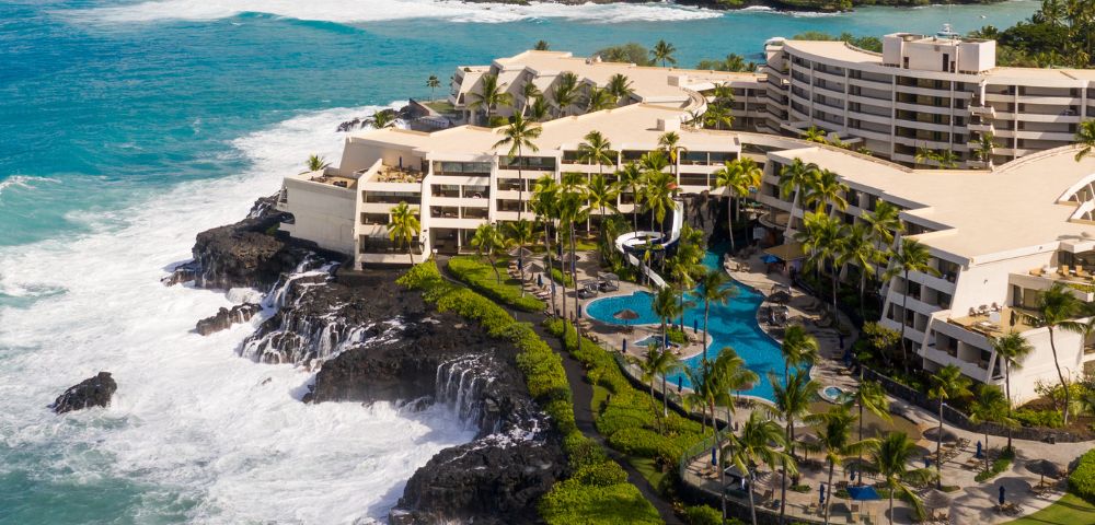 Aerial view of an oceanfront resort with cascading waves, lush palm trees, and a curving pool. Sunlit buildings create a serene, luxurious tropical setting.