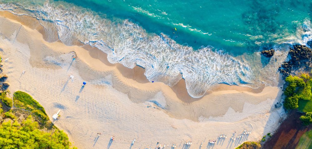 Aerial view of a tranquil beach with gentle waves lapping onto golden sand. Sparse umbrellas and sunbathers create a serene, relaxing atmosphere.