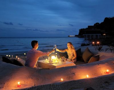 A couple enjoys a romantic candlelit dinner on a secluded beach at dusk. They sit by a table with wine glasses, surrounded by sand, cushions, and glowing candles.
