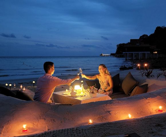 A couple enjoys a romantic candlelit dinner on a secluded beach at dusk. They sit by a table with wine glasses, surrounded by sand, cushions, and glowing candles.