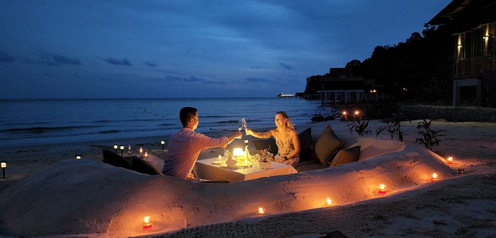 A couple enjoys a romantic candlelit dinner on a secluded beach at dusk. They sit by a table with wine glasses, surrounded by sand, cushions, and glowing candles.