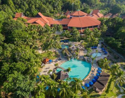 Aerial view of a tropical resort with a large, turquoise pool surrounded by palm trees and deck chairs with blue umbrellas. Red-roofed buildings in the background.