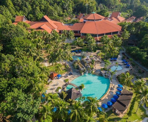 Aerial view of a tropical resort with a large, turquoise pool surrounded by palm trees and deck chairs with blue umbrellas. Red-roofed buildings in the background.