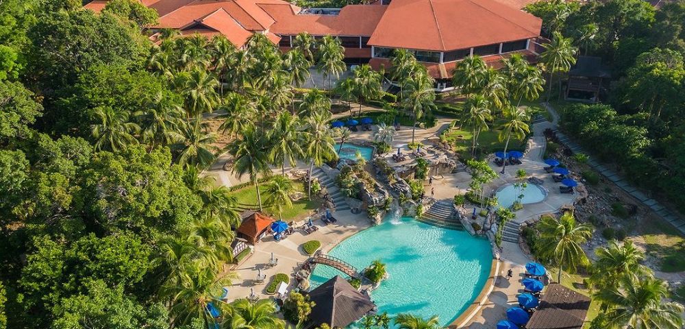 Aerial view of a tropical resort with a large, turquoise pool surrounded by palm trees and deck chairs with blue umbrellas. Red-roofed buildings in the background.