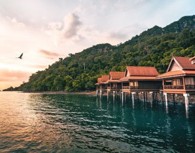 Coastal villas on stilts line a serene blue-green bay, backed by lush green hills. A bird soars above in the pink-hued, tranquil sunset sky.