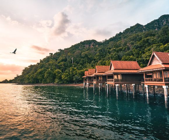 Coastal villas on stilts line a serene blue-green bay, backed by lush green hills. A bird soars above in the pink-hued, tranquil sunset sky.