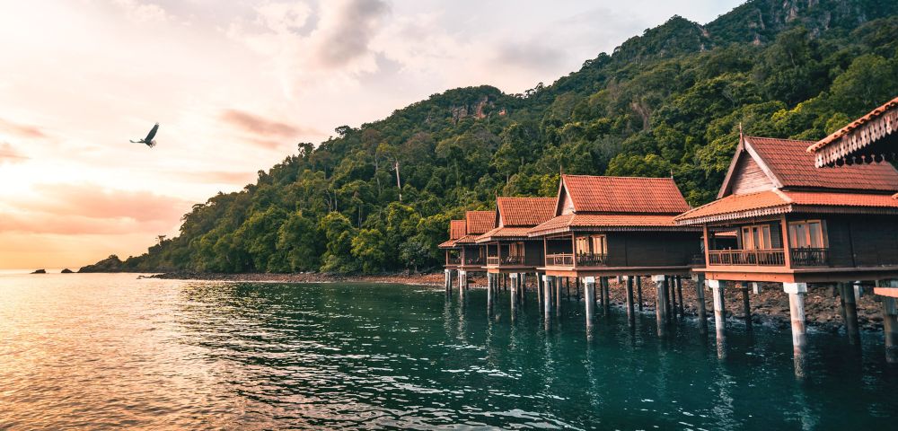 Coastal villas on stilts line a serene blue-green bay, backed by lush green hills. A bird soars above in the pink-hued, tranquil sunset sky.