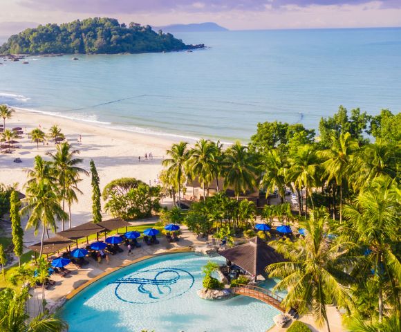 Tropical beach scene with blue ocean and lush greenery. A pool with a decorative design is surrounded by palm trees, sun loungers, and umbrellas.