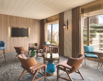 Modern lounge with wooden walls, brown leather chairs, small tables, a wall-mounted TV, and large windows overlooking a cityscape, creating an inviting atmosphere.