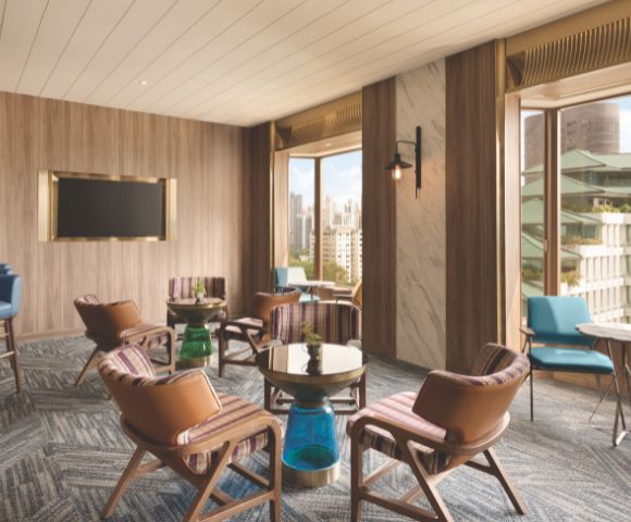 Modern lounge with wooden walls, brown leather chairs, small tables, a wall-mounted TV, and large windows overlooking a cityscape, creating an inviting atmosphere.