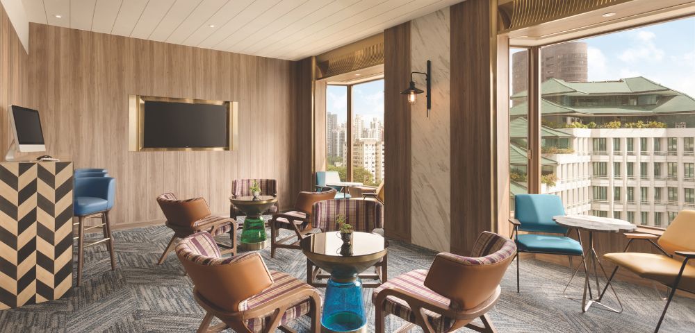 Modern lounge with wooden walls, brown leather chairs, small tables, a wall-mounted TV, and large windows overlooking a cityscape, creating an inviting atmosphere.