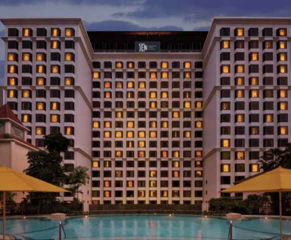 Tall hotel building at dusk with lit windows, reflecting a warm ambiance. A tranquil pool in the foreground is surrounded by yellow umbrellas.