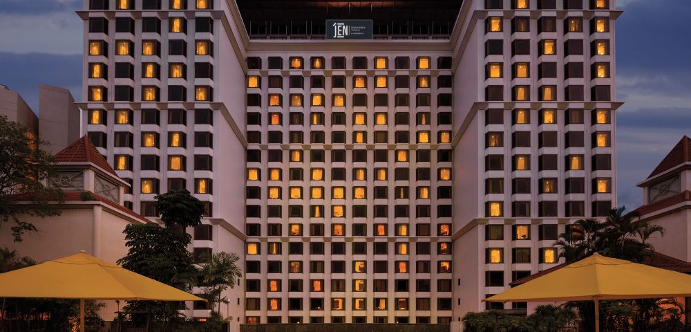 Tall hotel building at dusk with lit windows, reflecting a warm ambiance. A tranquil pool in the foreground is surrounded by yellow umbrellas.