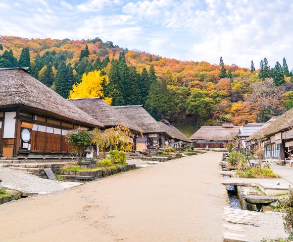 A traditional Japanese village with thatched-roof houses lines a dirt path. Autumn leaves cover the hillside, with vibrant yellows and oranges. Quiet, serene setting.