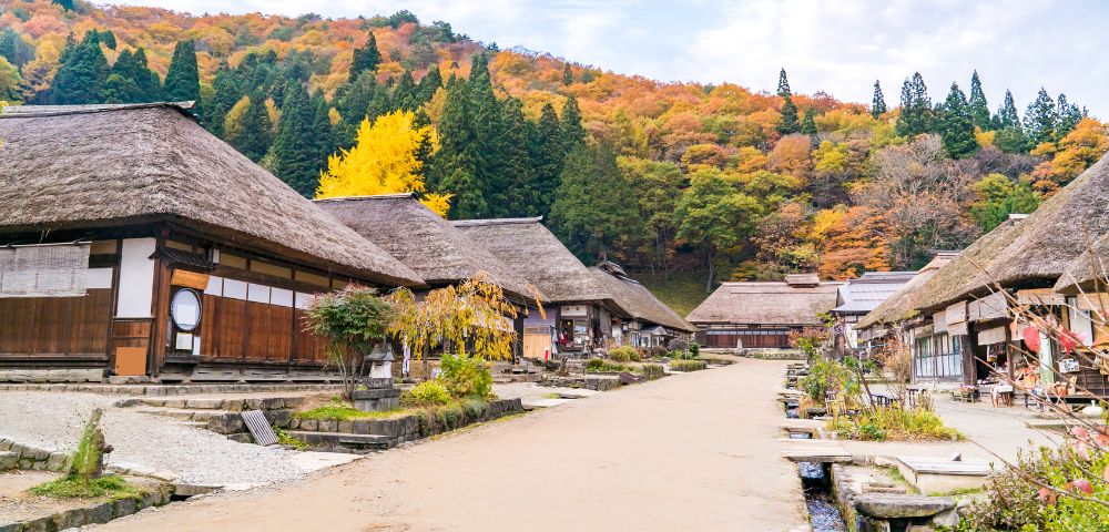 A traditional Japanese village with thatched-roof houses lines a dirt path. Autumn leaves cover the hillside, with vibrant yellows and oranges. Quiet, serene setting.