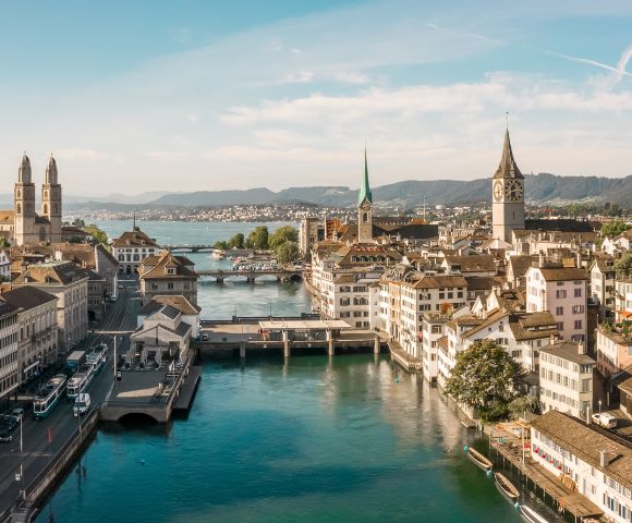 Aerial view of Zurich, Switzerland, showcasing historic buildings, church steeples, and a serene river under a bright blue sky. Calm and picturesque.