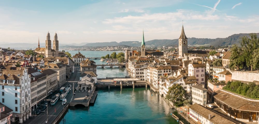 Aerial view of Zurich, Switzerland, showcasing historic buildings, church steeples, and a serene river under a bright blue sky. Calm and picturesque.