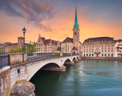 Scenic view of a historic bridge and church with a tall spire in Zurich at sunset. The sky is painted in warm hues, reflecting on the tranquil river.