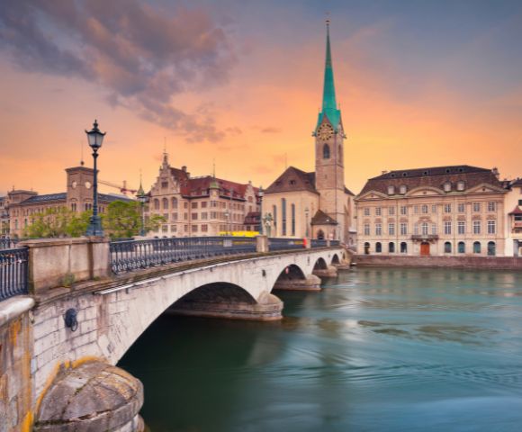 Scenic view of a historic bridge and church with a tall spire in Zurich at sunset. The sky is painted in warm hues, reflecting on the tranquil river.