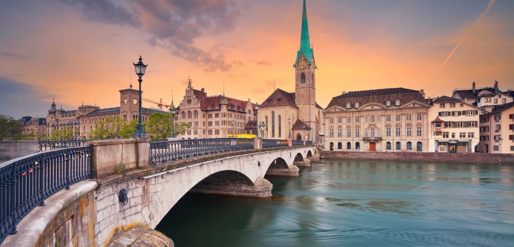 Scenic view of a historic bridge and church with a tall spire in Zurich at sunset. The sky is painted in warm hues, reflecting on the tranquil river.