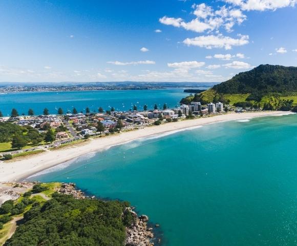 Aerial view of a coastal landscape with a turquoise bay, sandy beach, and lush green mountain. Blue sky with scattered clouds adds to the serene vibe.