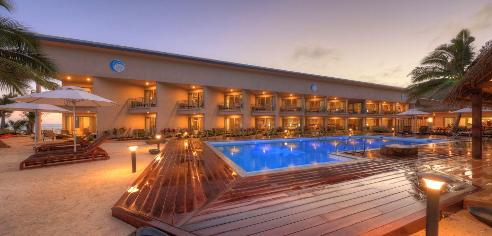 Elegant resort at sunset with a glowing blue pool, surrounded by palm trees and loungers. The tranquil ambiance suggests relaxation and luxury.