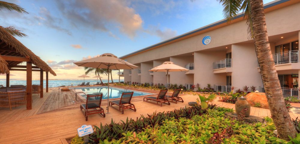 Resort poolside area with lounge chairs and umbrellas, surrounded by lush greenery and palm trees at sunset, evoking a peaceful, tropical ambiance.