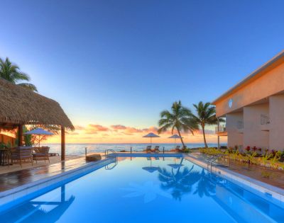 Luxurious seaside resort with a tranquil pool reflecting a stunning sunset. Palm trees, thatched roof shelter, and loungers evoke a serene, tropical paradise.