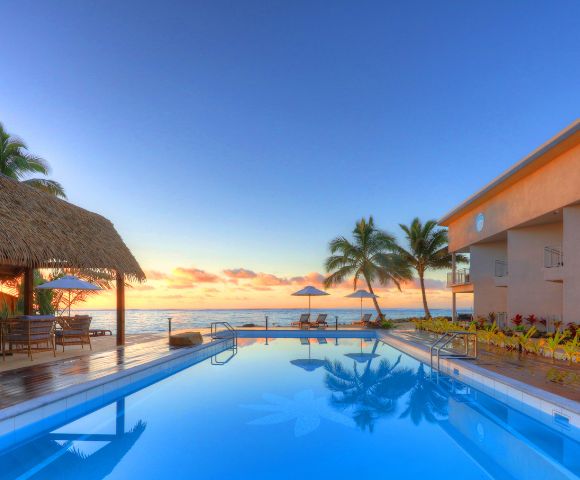 Luxurious seaside resort with a tranquil pool reflecting a stunning sunset. Palm trees, thatched roof shelter, and loungers evoke a serene, tropical paradise.