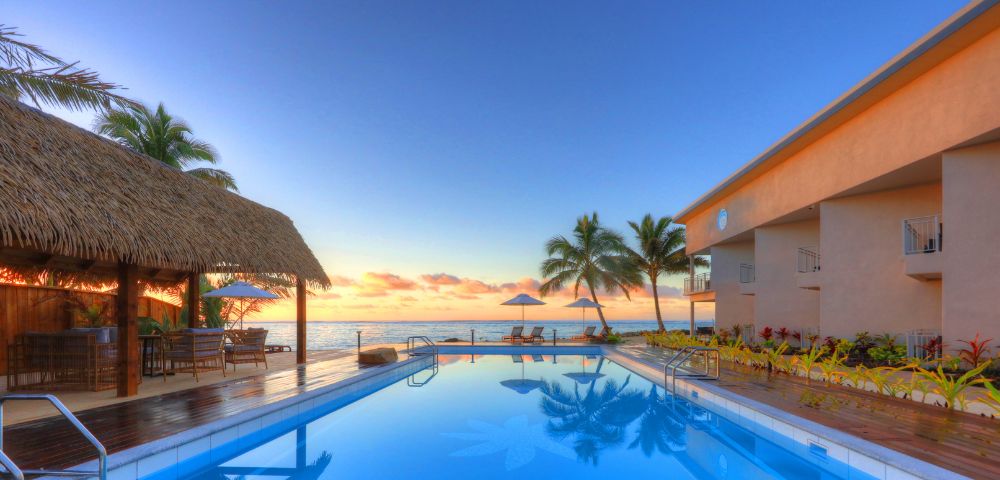 Luxurious seaside resort with a tranquil pool reflecting a stunning sunset. Palm trees, thatched roof shelter, and loungers evoke a serene, tropical paradise.